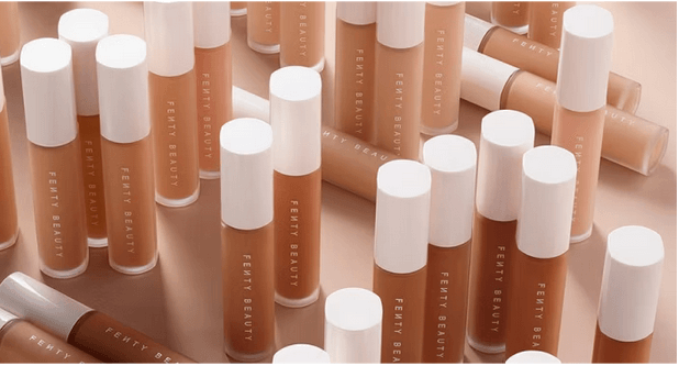 A collection of Fenty Beauty foundation sticks in various shades of beige to brown, arranged aesthetically against a matching beige background. The variety of shades highlights the brand’s focus on inclusivity for different skin tones.