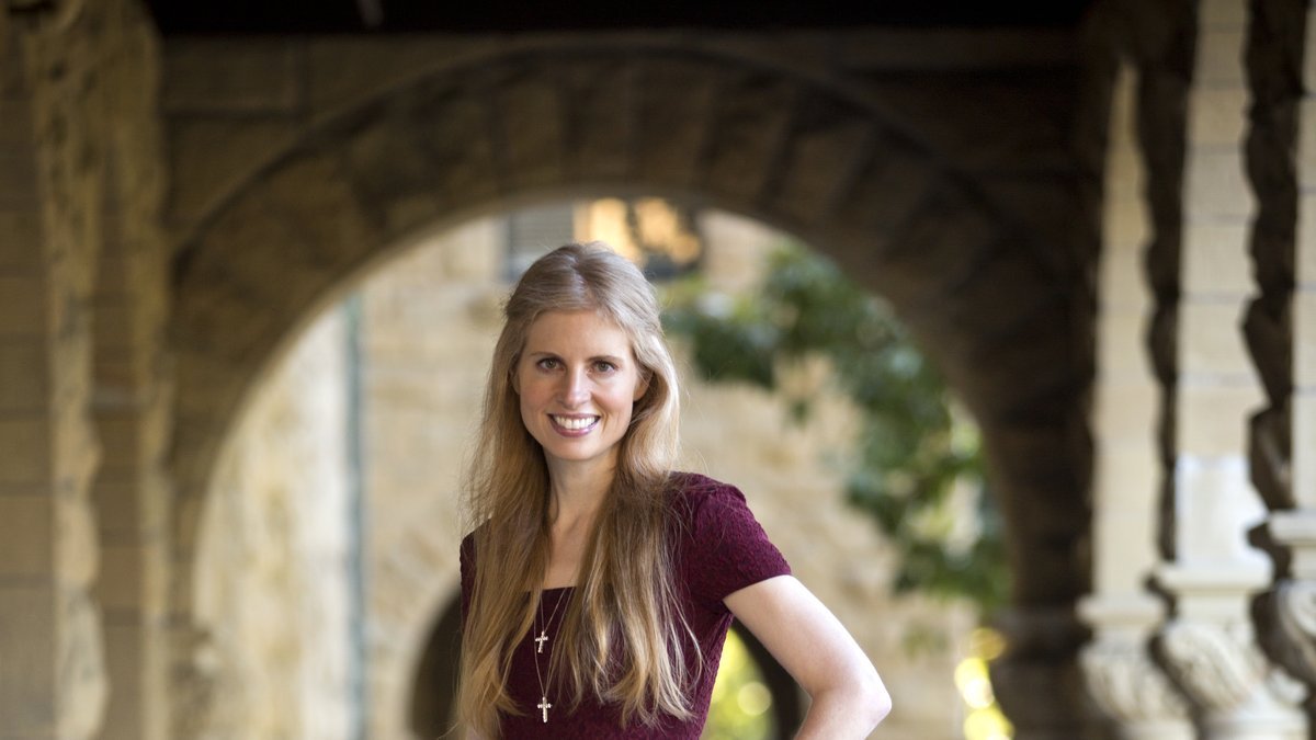 The image shows Laura Arillaga, wife of tech entrepreneur Marc Andreessen. She is smiling warmly, standing outdoors in front of a stone archway, with a background of greenery and historical architecture. Laura is dressed in a maroon dress and appears to be in a serene, elegant setting