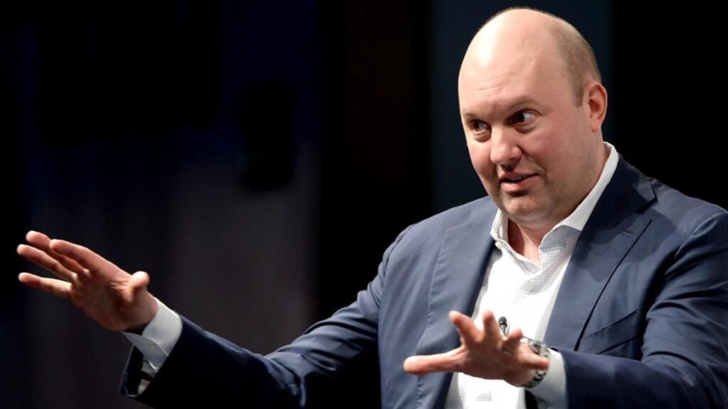 The image shows Marc Andreessen, a prominent tech entrepreneur, speaking or explaining something. He is dressed in a blue suit and a white shirt, with his hands raised in an expressive gesture. His focus is intense, and he appears to be engaged in a detailed conversation. The background is blurred, with a dark backdrop that helps highlight his presence in the moment