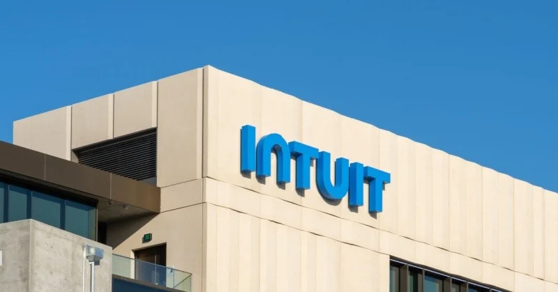 This image shows the Intuit logo on the side of a modern building. The logo is blue and white, with the word "Intuit" spelled out in large, bold letters. The building is tan and has a flat roof with a small vent. The sky is blue and clear.