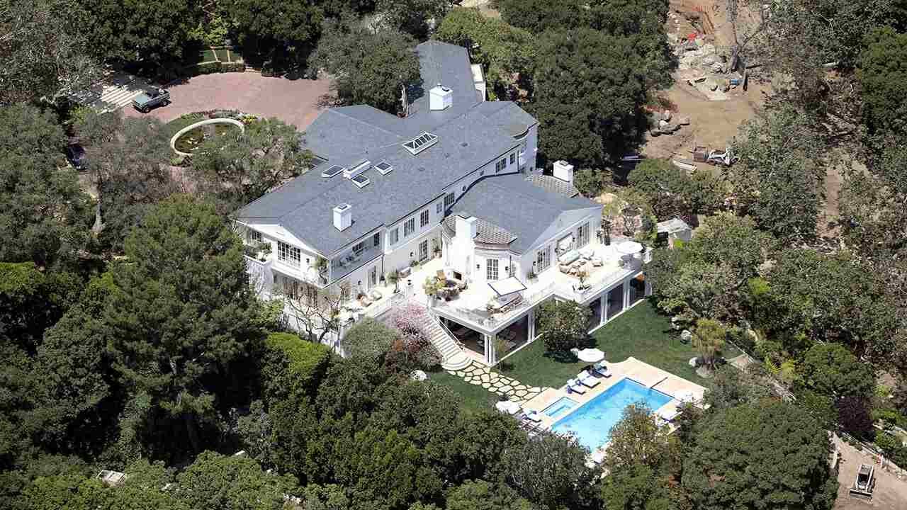 This image shows an aerial view of a large, luxurious Jeff Bezos mansion. The house is white with a gray roof and has multiple levels and wings. There is a swimming pool and a large lawn area in front of the house. The property is surrounded by lush green trees.