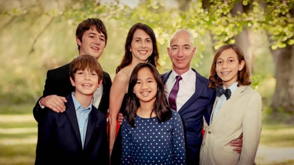 Jeff Bezos smiling alongside his family, including four children and a woman, all standing outdoors in a lush, sunlit setting with greenery in the background.