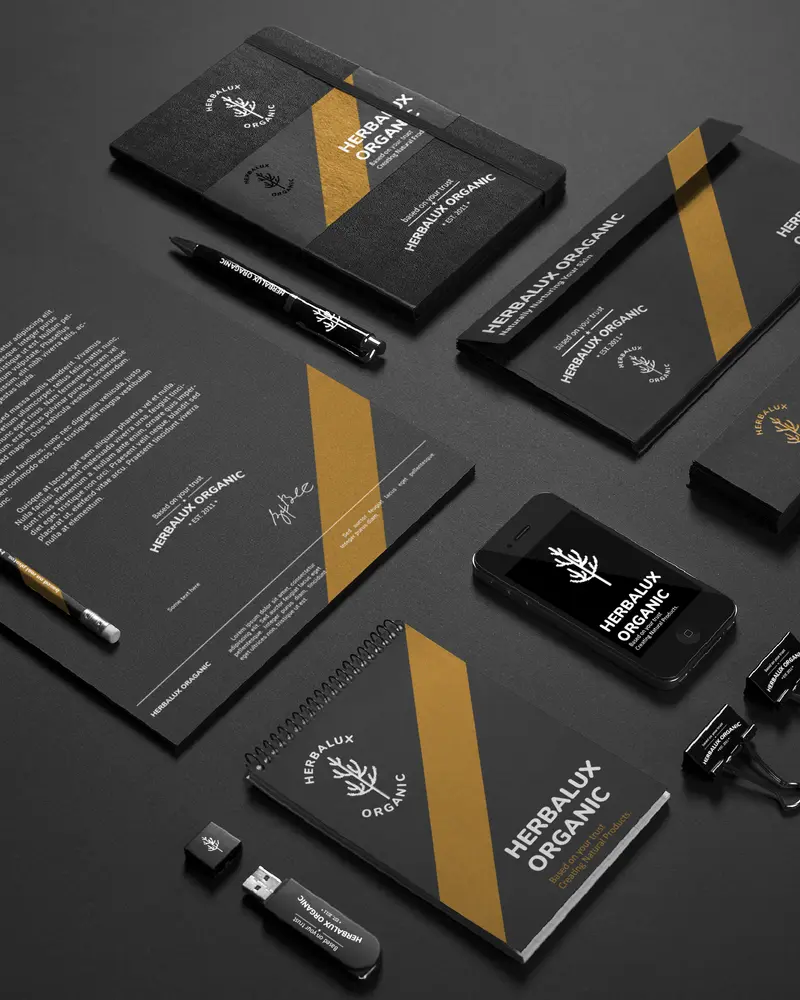 Branded corporate materials for HERBALUX ORGANIC, including business cards, a notebook, a pen, an envelope, a USB stick, and a mobile phone, all featuring a stag’s head logo with antlers in white on black background and gold accents.